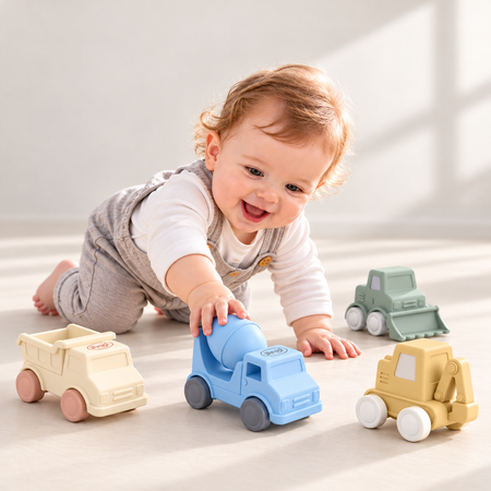 A baby crawling on a light floor and reaching for a blue toy cement mixer truck, with other toy construction vehicles nearby.