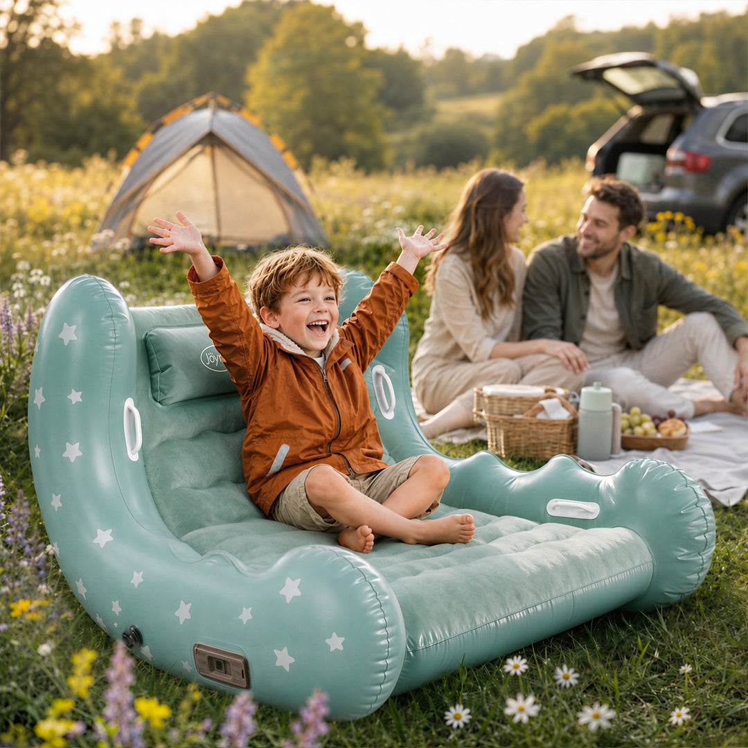 A child sits happily on a large mint green inflatable lounge chair with white star patterns in a grassy field. In the background, a couple sits on a picnic blanket near an open car trunk, with a camping tent visible in the distance.