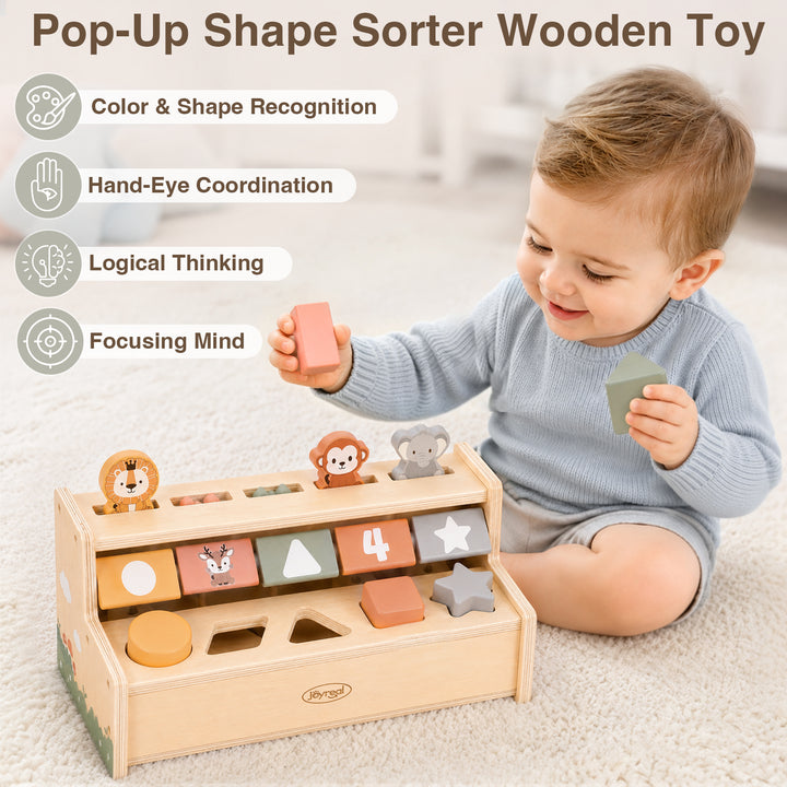 A toddler sits on a cream carpet playing with a wooden pop-up shape sorter toy. The toy has a natural wood frame with colorful geometric blocks and animal cutouts.